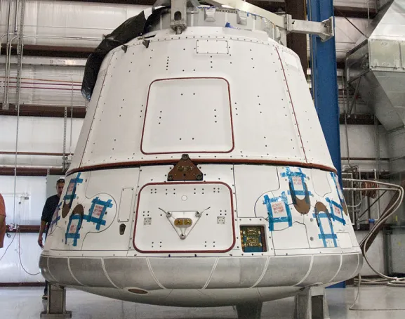 Photo du vaisseau cargo SpaceX Dragon, un grand conteneur blanc en métal. Il a la forme d’une pyramide ronde sans sommet et est posé sur trois pattes dans un genre de grand entrepôt.