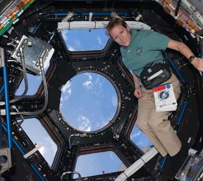Photo de Kathryn Hire, en apesanteur dans une coupole d’observation de la Station spatiale internationale. Elle porte à la taille un sac banane auquel est attaché un manuel quelconque. On voit l’extérieur de la Station par les hublots derrière elle.