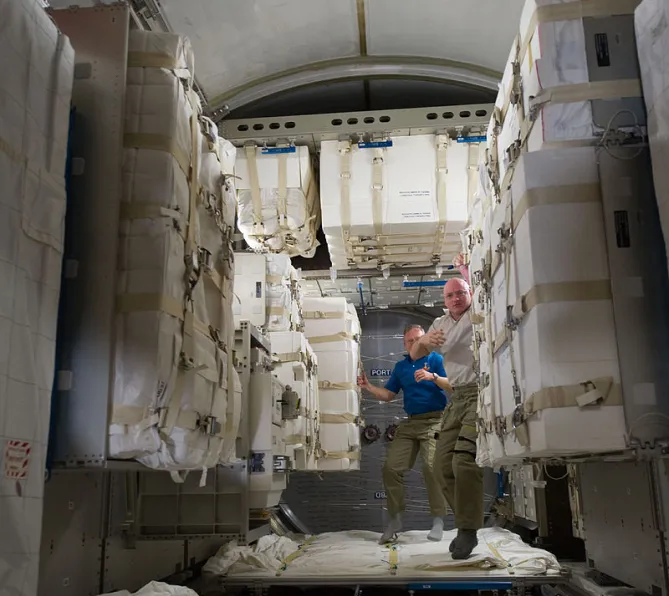 Photo de deux hommes qui circulent en apesanteur dans un couloir du module Léonardo de la Station spatiale internationale. De chaque côté d’eux et au-dessus d’eux, une multitude de paquets emballés sont stockés le long des murs.