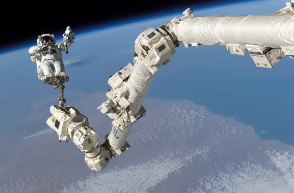Photo du Canadarm2, vu de la Station spatiale internationale avec la Terre en arrière-plan. Un astronaute en combinaison est accroché debout au bout du bras robotique articulé, blanc.