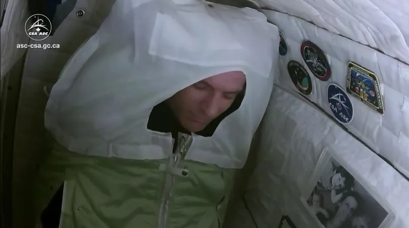 Photo de l’astronaute David Saint-Jacques. Il et emmailloté, les yeux fermés, dans un sac de couchage coussiné dans une petite cabine de repos.