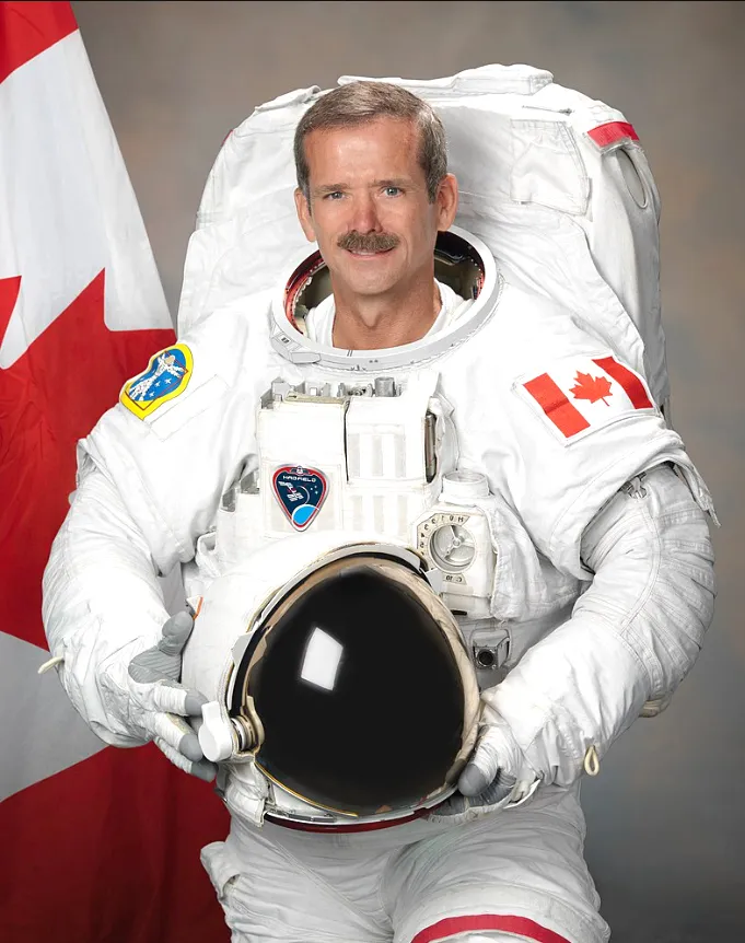 Photo de Chris Hadfield, en habit d’astronaute, se tenant debout devant le drapeau du Canada.