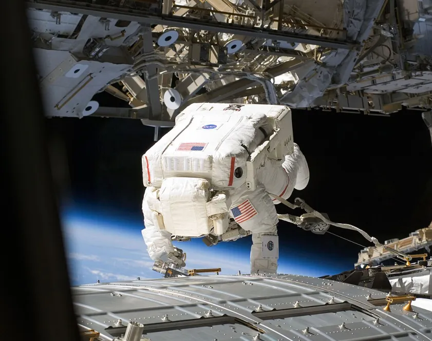 Photo d’un astronaute en combinaison incluant le casque qui travaille sur le module Kibo, à l’extérieur de la Station spatiale internationale à laquelle il est attaché par un long câble.
